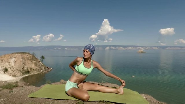Young professional yoga woman practices yoga moves outdoors. blue lake baikal on the background
