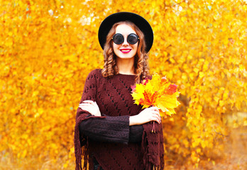 Autumn fashion smiling woman in black round hat, knitted poncho on a sunny yellow leaves background