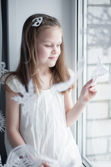 Fototapeta na wymiar girl in white dress sits on window sill among snowflakes and looks out the window