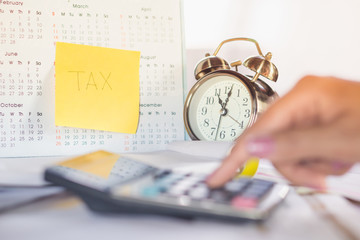tax word note on calendar, alarm clock and calculator on table with blur woman hand calculating in...