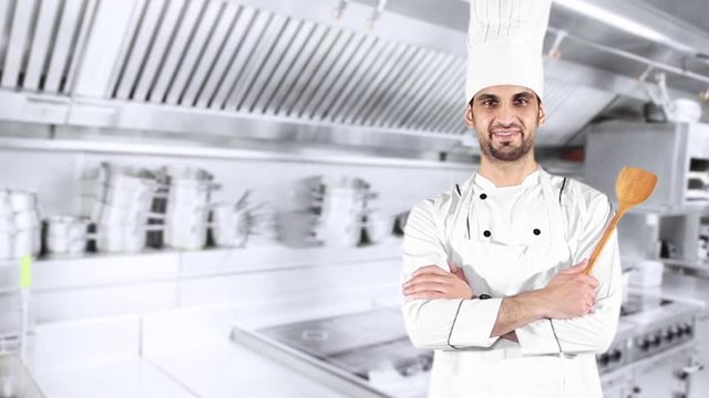 Male Caucasian chef holding a wooden spatula and smiling at the camera while crossed hands and standing in the kitchen