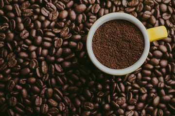 Small, yellow espresso cup filled with coffee powder with coffee beans all around. 
