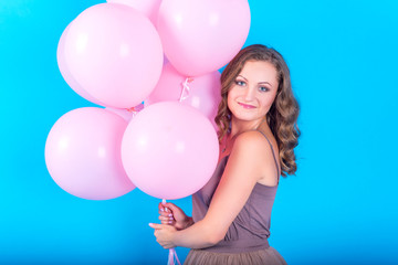 Happy smiling young woman having fun with pink helium air balloons over blue background. Girl with curly hair in dress holding pink balloons. Beauty,  holidays, birthday, valentine, fashion concept