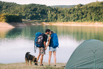 Obraz na płótnie Canvas Hiking couple. Young couple with backpacks and dog kissing beside lake 