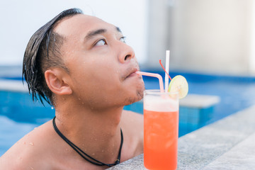 Portrait of a handsome man resting at the edge of a swimming pool. Tropical drink with drinking straw and paper umbrella next to him. Summer vacation concept.