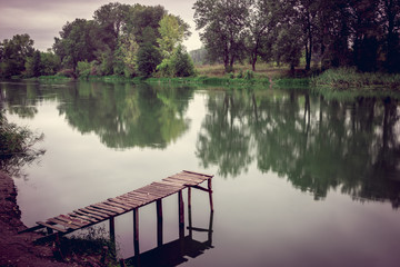 Pier on a calm river in the summer. Wooden pier bridge in the morning. Place for fishing in the river. Toned, style photo.