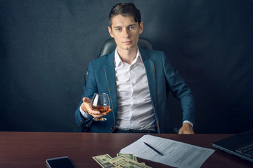 Man in suit sitting in chair. Boss drink alcohol in the glass and enjoys the success. Concept of a successful business