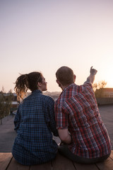 Romantic date on roof. Couple watching sunset. Unrecognizable modern people in focus on foreground, male pointing at sky. Pleasant pastime together, urban background with free space