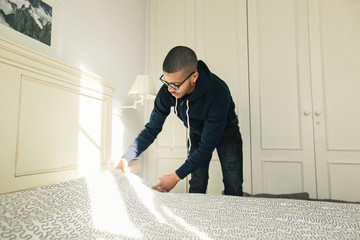 Latin man making bed at home.
