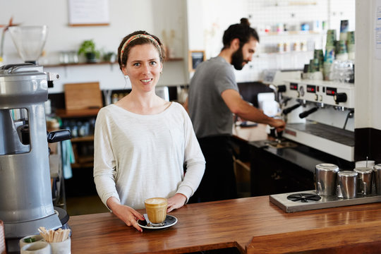 Young woman offering latte while her colleague working behind