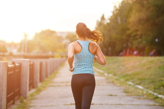 Young smiling sporty woman running in park in the morning. Fitness girl jogging in park. Rear view of sporty girl running on embankment