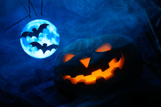 Scary Halloween pumpkin with a glowing face and bats on background of the full moon in the fog