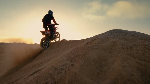 Following Shot of the Professional Motocross Motorcycle Rider Driving Over the Dune and Further Down the Off-Road Track. It's Sunset and Track is Covered with Smoke/ Mist. 4K UHD.