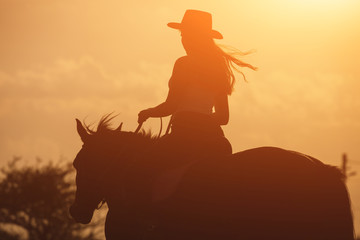 Sunset silhouette of young cowgirl riding her horse - obrazy, fototapety, plakaty