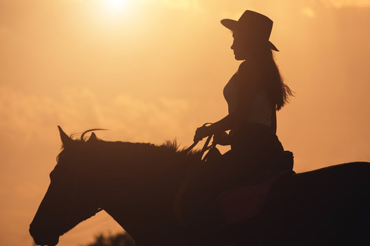 Fototapeta Sunset silhouette of young cowgirl riding her horse