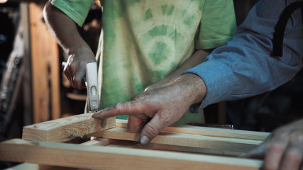 Unrecognizable grandson helps nailing wood to grandfather carpenter.