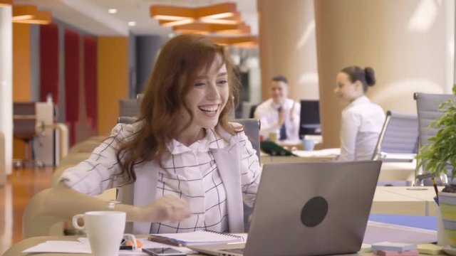 young happy woman shouts and laughs when see good news of success in computer during working day with papers sitting at table in co working colorful business office space