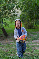 A little boy in jeans clothes plays with a bright ball in football. A funny kid in a hood holds a bright round ball. Football a second before kicking the ball.