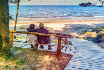 Happy elderly couple resting on the wooden bench and  looking at the distance of the Baltic Sea. Image slightly toned for inspiration of retro style. - obrazy, fototapety, plakaty