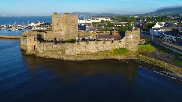 Medieval Norman Castle in Carrickfergus near Belfast, Northern Ireland,  in sunrise light. Aerial flyby video with marina, yachts, parking, town and far view of Belfast in the background