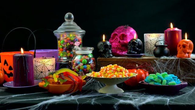 4k Happy Halloween trick or treat party table with bowls and apothecary jars of candy with skull candles against a black background, full table static.