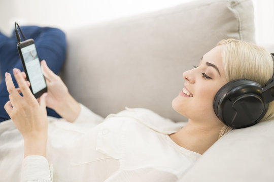 The happy woman with an earphones and phone lay on the sofa