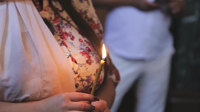 Young woman is holding burning candle while standing in church indoors. Female holds lighted candle in arms while on Sunday worship service in temple. Parishioner dressed in colorful clothes keep