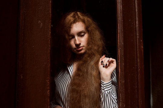 Young sad red haired model with long hair standing behind a closed glass door. Raindrops on the glass