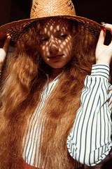 Fototapeta premium Elegant young red haired model in straw hat with shadow on her face