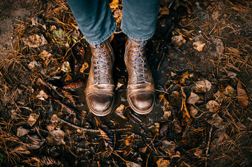 Closeup of man`s legs in fashinable trendy italian hipster vintage old scratched boots standing in puddle. Wet and raw autumn. Fall colors. Orange leaves, pine cones in water. Odd bizarre concept.