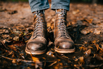 Closeup of man`s legs in fashinable trendy italian hipster vintage old scratched boots standing in puddle. Wet and raw autumn. Fall colors. Orange leaves, pine cones in water. Odd bizarre concept.