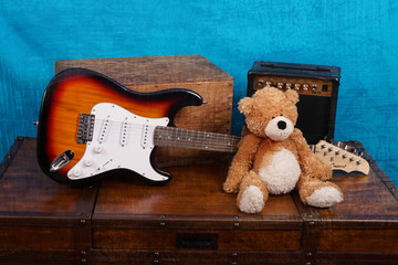 brown and white teddy bear sitting by an electric guitar against a blue background.