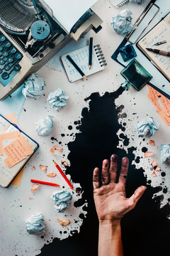 Writer workplace with spilled ink, crumpled paper, scattered letters, stationery and a typewriter. Ink leaving stains on an open palm. Space for creative lettering.