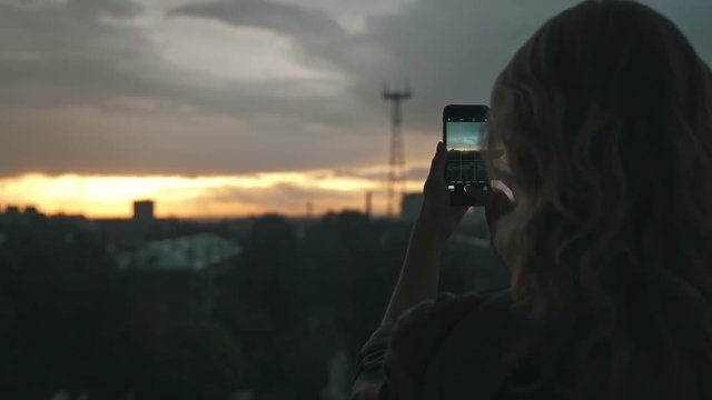 Female silhouette: girl in plaid photographing sunset on a smartphone. Beautiful fiery sunset: women's hands hold mobile phone, photographing. Girl is not visible, only back and shoulders.