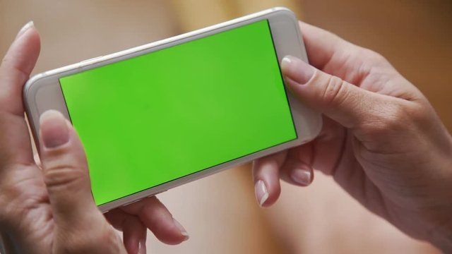 Female hands holding a white smartphone with a green screen on the background of a wooden floor. Modern technologies and gadgets: using the phone. The phone is in a horizontal position.