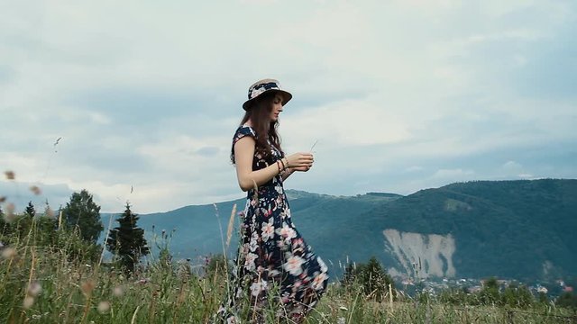 Young Pretty Woman Wearing Hat and Dress is Picking a Flowers on Mountains Background.