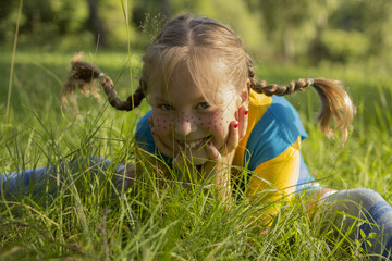 smiling blond girl with funny pigtail sits in grass. She is wearing a yellow T-shirt and a blue sarafan, and she has freckles on her nose.
