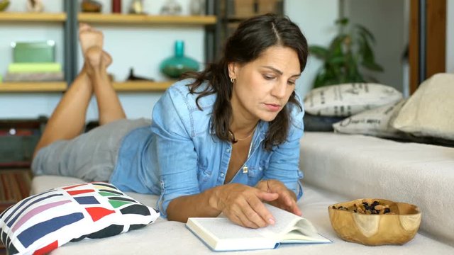 Woman in denim shirt looks bored while reading book on the sofa
