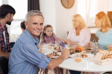 A man in a blue shirt is sitting at a table and posing against the background of his family, who is sitting and eating