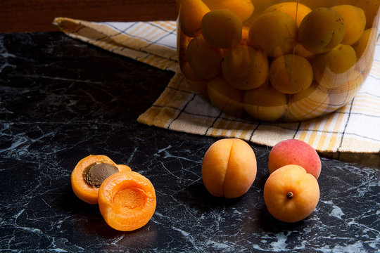 Juicy harvested apricots and big glass jar with canned apricot compote on dark marble..