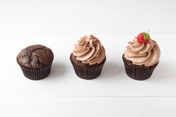 Cupcakes with whipped chocolate cream, decorated fresh strawberry on white wooden table. Picture for a menu or a confectionery catalog, showing different stages of creation.
