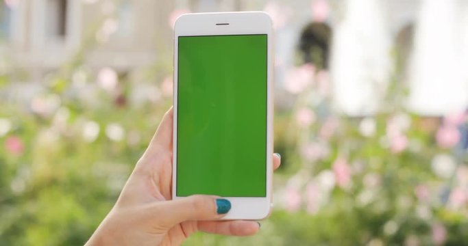 Woman hand holding using mobile smart phone blurred background garden flowers green screen chromakey tapping touch pointing finger display closeup outside modern lifestyle trendy app 3g online gadget