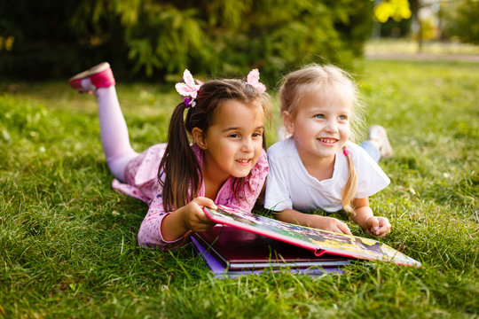 Two young girls lying on the grass outdoors reading separate books. These siblings are enjoying time together as family during summer