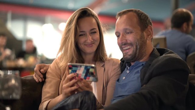 Happy couple talking and watching movie on smartphone in cafe
