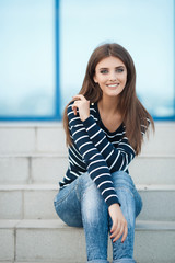 Young beautiful woman brunette with long straight hair and gray-green eyes,dressed in a striped t-shirt and blue jeans,spends time in the city in the spring,sitting on the steps,one in the fresh air