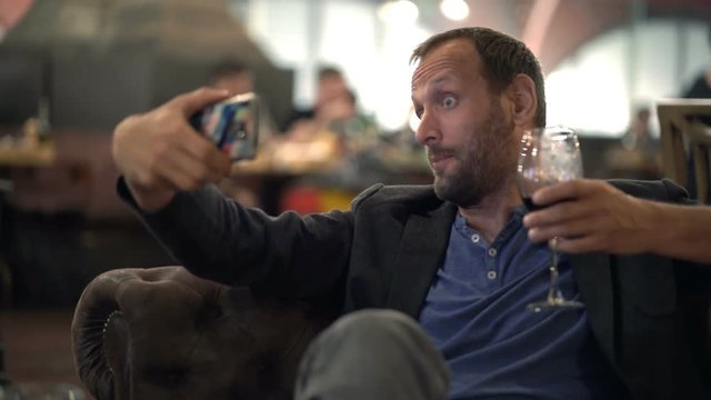 Happy man with wine taking selfie photo with cellphone on sofa in cafe
