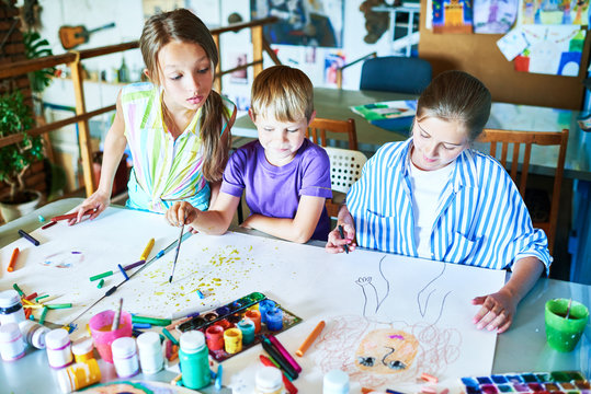 High angle  portrait of three little kids painting pictures in art class sitting at tables with art supplies, pencils and paints