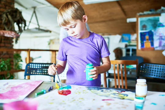 Portrait of blonde little boy painting pictures  carefully, standing at table in art studio