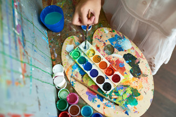Above view image of watercolor paints tray on top of messy palette with color blots used by unrecognizable child painting on easel during art class