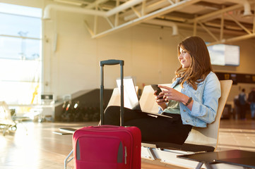Airport Young female passenger on smart phone and laptop sitting in terminal hall while waiting for...